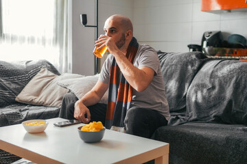 Young man drinking beer while watching sports at home with a blue and red sports scarf
