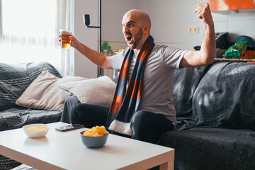 Young man celebrating while watching sports on the sofa with a beer