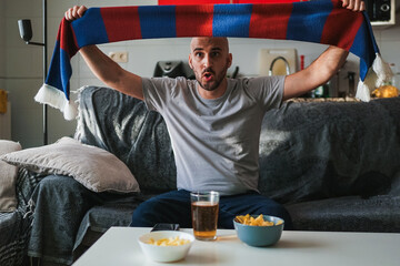 Young man cheering on his team in the sofa with a scarf