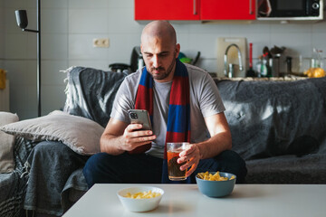 Young man looking at the phone while watching sports on the sofa - obrazy, fototapety, plakaty