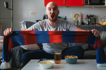 Young man screaming with his team's scarf watching sports on the sofa