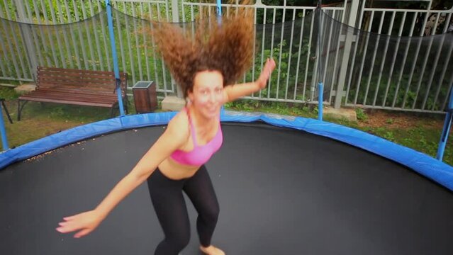 Woman in sports clothes jumps on round trampoline near fence