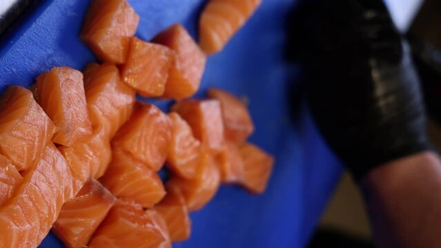 chef man hand is cutting salmon on blue board in restaurant, preparing dish for catering.cubes of fish with green spinach over ging in grill oven. close up from kitchen 4k