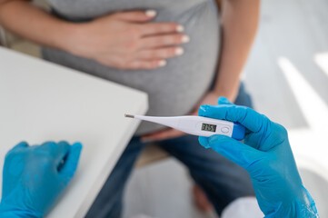 Pregnant woman with fever at doctor's appointment. Therapist holds an electronic thermometer with a temperature of 37.5. 