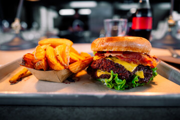 A delicious hamburger with two beef patties, bacon, lettuce, cheese and brioche bun, accompanied by chunky waffle cut fries served on a tray with restaurant kitchen, glass & drink in bokeh background