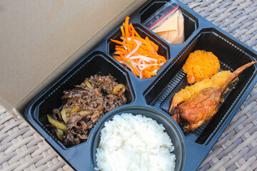Indonesian Bento box containing rice, beef teriyaki, salad and dressing, and fried snacks.