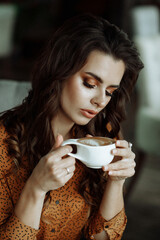 A serene Caucasian woman with wavy hair, in a polka-dotted blouse, savoring her coffee in a cozy, softly lit interior. Front view. Concept for a tranquil morning routine.
