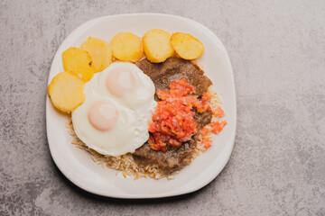 Top view of authentic, traditional Bolivian meal: Silpancho, made of beef, potatoes, rice, fried eggs, tomato and onion salad. close-up on white plate on gray background.copy space