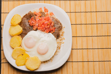 Top view of authentic, traditional Bolivian meal: Silpancho, made of beef, potatoes, rice, fried eggs, tomato and onion salad. close-up on white plate  on bamboo tablecloth.copy space