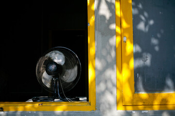 Black electric fan in the yellow window with shadow of leaves in the summer day