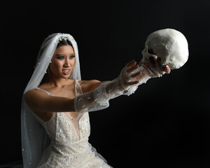 Close up portrait of beautiful model wearing white wedding gown and ghostly flowing veil like a shroud.  Holding a skull skeleton.  isolated on a dark studio background.