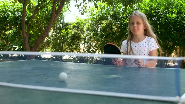 Pretty emotion girl play table tennis on green bushes background. Blond child player hit lightweight ping-pong ball back across hard table divided by tennis net use small red rackets. Real people