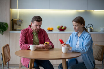 Wife and husband with smartphone addiction sitting in kitchen, scrolling web pages, ignoring each other. Married couple dependent on internet, social networks. Absence of communication in family life