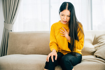 Illustrating a heart attack, Asian woman experiences chest pain needing immediate medical help while on a sofa. Reflecting heartache discomfort and the urgency for emergency aid. - obrazy, fototapety, plakaty