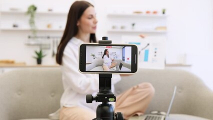Concept of technology and blogging. Young caucasian brunette financier woman filming herself on cell phone which fixed on tripod and showing financial statistics to subscribers in living room.