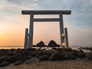 A serene and tranquil scene of a beach at sunset, with a large, white torii gate on the rocky shore.