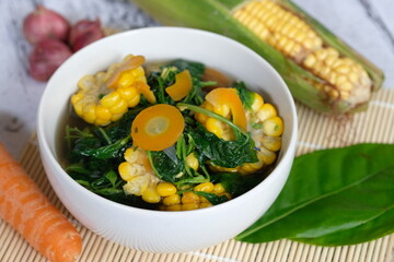Sayur Bening Bayam is spinach clear soup, made of fresh spinach, carrot, corn, red onion, bay leaf. This soup served in white bowl. Indonesian food. Top view, flat lay, wooden table. 
