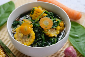 Sayur Bening Bayam is spinach clear soup, made of fresh spinach, carrot, corn, red onion, bay leaf. This soup served in white bowl. Indonesian food. Top view, flat lay, wooden table. 
