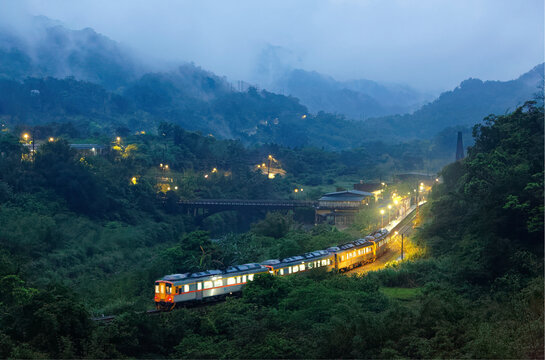 A local train travels in a beautiful valley surrounded by misty mountains on a foggy spring evening, lights of a highway glow in dusk and a bridge spans the stream near Wanggu Station, Taipei, Taiwan