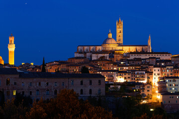 Fototapeta premium Night scene of Siena, a medieval town on a hilltop in Tuscany, Italy, with a view of architectural landmarks (Dome & Bell Tower of Siena Cathedral and Mangia Tower) lighted up in blue evening twilight