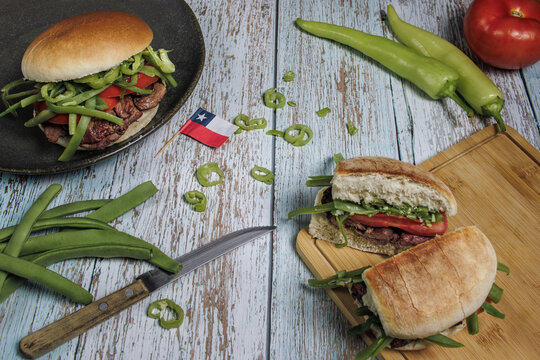 Chacarero Aerial view with chili, green beans, meat, and tomato on a cutting board, round plate on a wooden table. Includes ingredients, knife, and Chilean flag. Typical food and drink of Chile