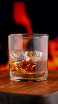 Glass of whisky with ice on wooden tabletop in front of fireplace, closeup shot