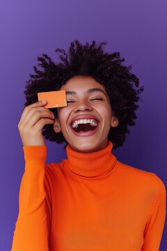 Vibrant and joyful woman in an orange turtleneck laughing and holding a credit card , against a purple background.