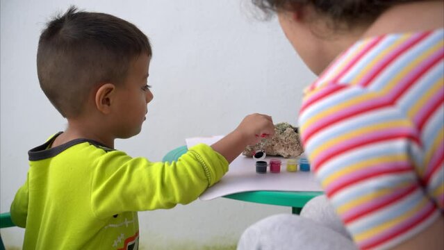 Slow motion of a young latin boy painting rocks with his aunt teacher instructor using small brushes