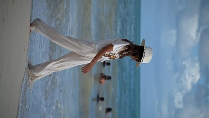 Vertical slow motion of a beautiful latin brunette woman walking on the beach wearing linen pants a white hat and yellow sunglasses enjoying the sea