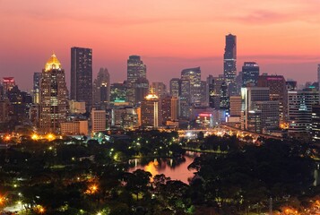 Urban skyline of Bangkok City at sunset, with famous landmark Mahanakhon Tower amid modern high rise buildings in background & the dramatic fiery sky reflected in a lake in the beautiful Lumphini park