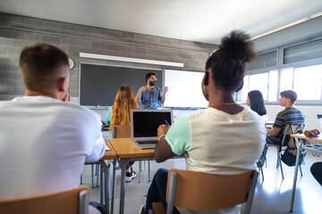 Rear view of multiracial group of teen high school students in classroom listen to male teacher...