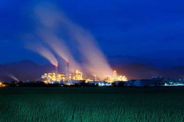 Fotobehang View of a chemical factory with smoking chimneys in the middle of a green farmland in the early morning twilight ~ Factory pipes polluting air in a silent morning, a serious environmental issue © AaronPlayStation
