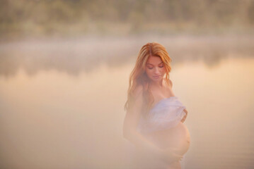 Fototapeta na wymiar Pregnant girl with long red hair by the river. At dawn. Fog near the water. Tenderness. Pregnancy. Future mom. Sunrise. Summer. Belly of a pregnant woman. Beautiful mother. Family. 9 months. Pending.