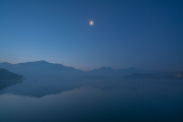 Fototapeta na wymiar A dawn at Sun Moon Lake in Nantou County, Taiwan. A beautiful morning with half moon over a lake in the mountains. Blue sky with reflection on the lake