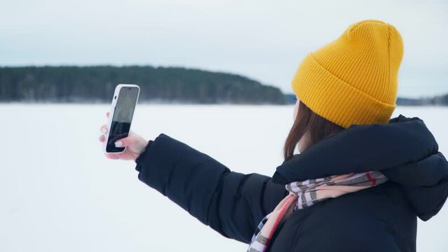 A beautiful young girl in a yellow cap takes a selfie on her phone. A girl takes a photo against the background of winter nature.