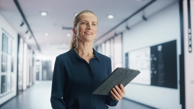 Adult Businesswoman Smiling While Checking Financial Documents on a Digital Tablet Computer. Portrait of a Successful Female Corporate Manager Walking in a Spacious Modern Office Building