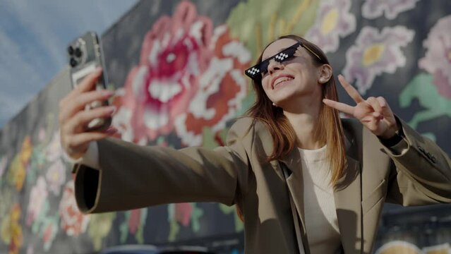 Woman in modern sunglasses poses for selfie against colourful wall with graffiti flowers. Young woman makes V sign with fingers to take photo on smartphone having fun. Cheerful way of life