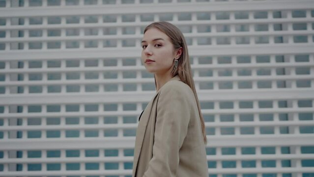Young stylish woman walks confidently past lacy net wall turning head to camera. Fashionable model stops to take position for photoshoot in street in day time. Photo shoot for magazine
