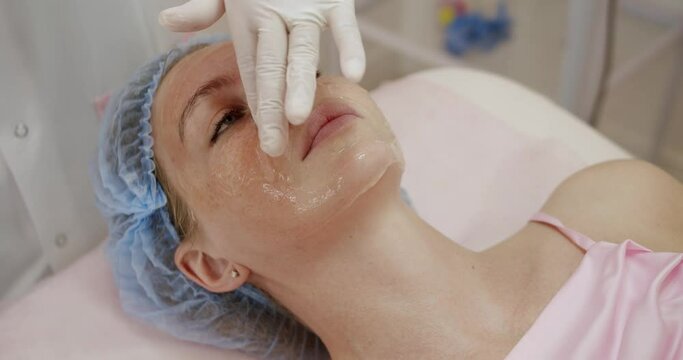 Woman lies on cosmetologist couch getting facial mask for rejuvenation treatment. Cosmetologist applies gel to female client face to cleanse skin prior to procedure. Cosmetic care