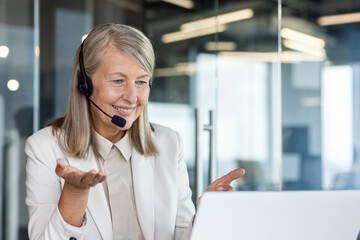 Positive mature woman dressed in stylish office outfit and headset looking at computer screen while...