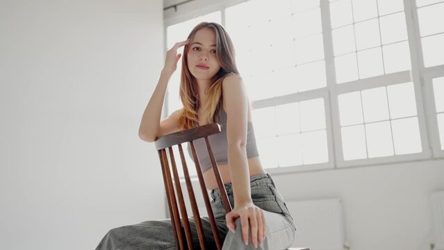 Young woman model leans on chair back at photo session in studio with panoramic windows. Lady with loose hair strikes best positions on chair for best photos. Model professional performance