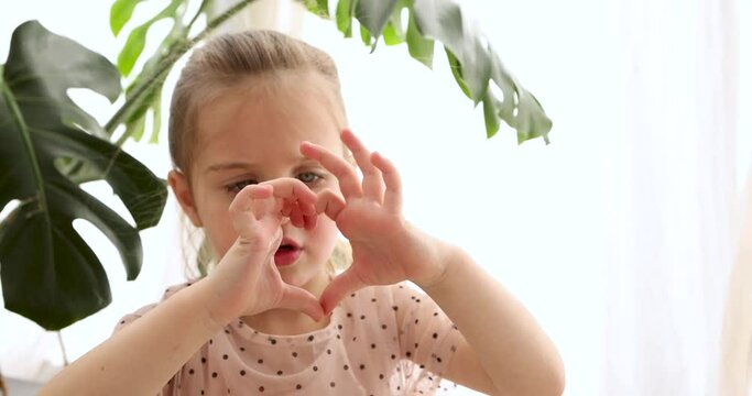 Cute little girl making hearts out of her hands, love, Valentine's day, showing a heart-shaped sign with her fingers. Portrait of small baby on white background with a house plant