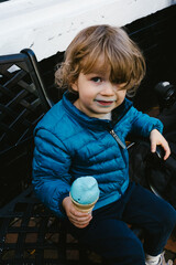 Little boy in blue coat eats blue ice cream on city bench