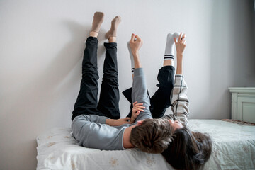 A young couple enjoys a relaxed, affectionate moment together while lying on a bed, with their legs playfully extended upwards against a white wall.