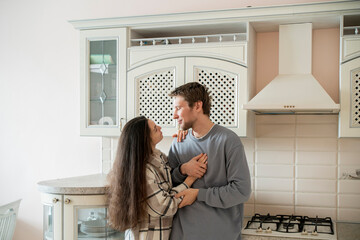 A couple stands close together in an affectionate embrace, exchanging loving looks in the comfort of their sunlit kitchen.