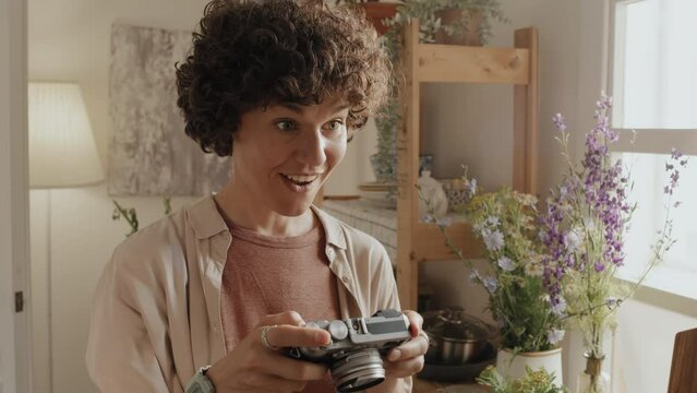 Creative Caucasian woman with curly hair holding camera standing in cozy kitchen taking photos of her family member