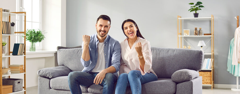 Excited couple celebrating success while sitting on sofa at home. Overjoyed happy young man and woman in casual wear raising clenched fists in excitement doing winner gesture
