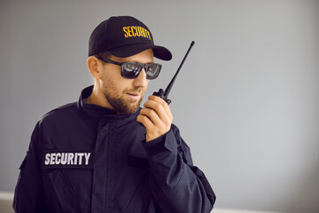 Security guard man in black uniform cap, jacket and sunglasses standing inside house and talking on modern portable wireless two way walkie talkie transceiver radio set device. Property safety concept