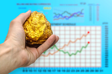 Gold nugget in a man’s hand against the backdrop of rising stock prices. Stock Exchange. Concept...