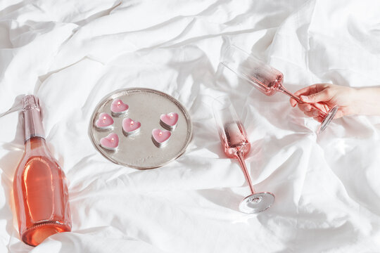 Woman hand holding shiny champagne glass, bottle of sparkling rose wine, pink candle hearts on bed cloth. Minimal lifestyle aesthetic photo, romance mood, Valentine's Day, romantic love concept